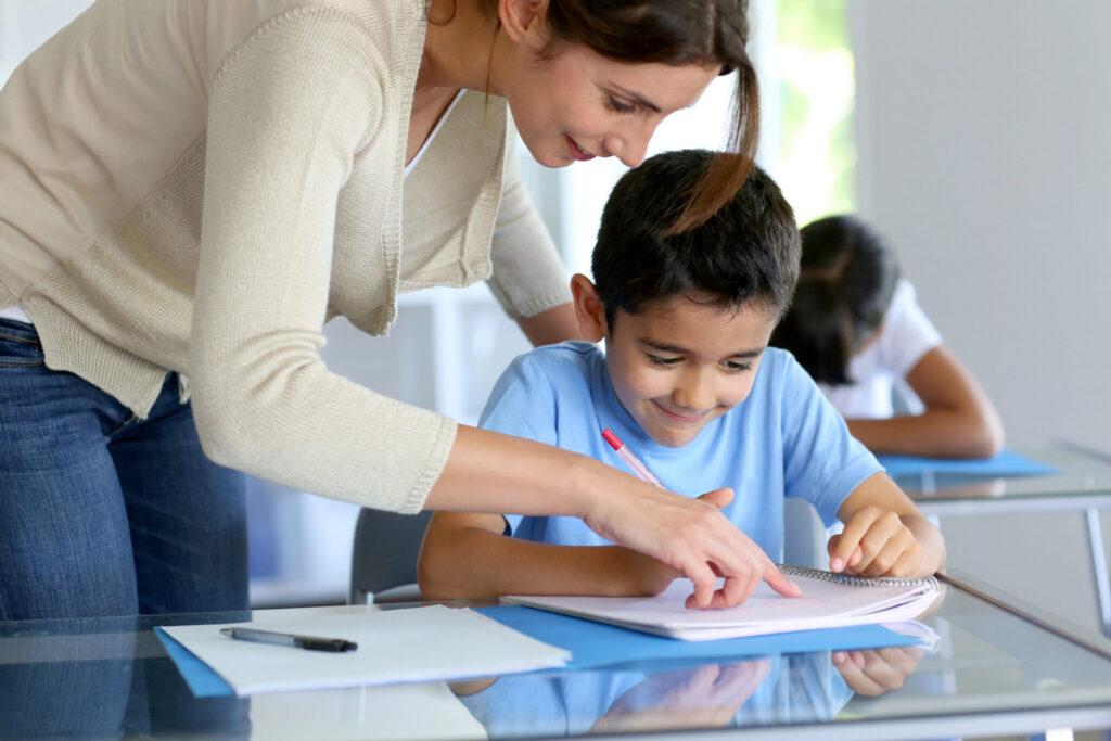 An adult helps a young boy with his schoolwork at a glass table in a classroom.