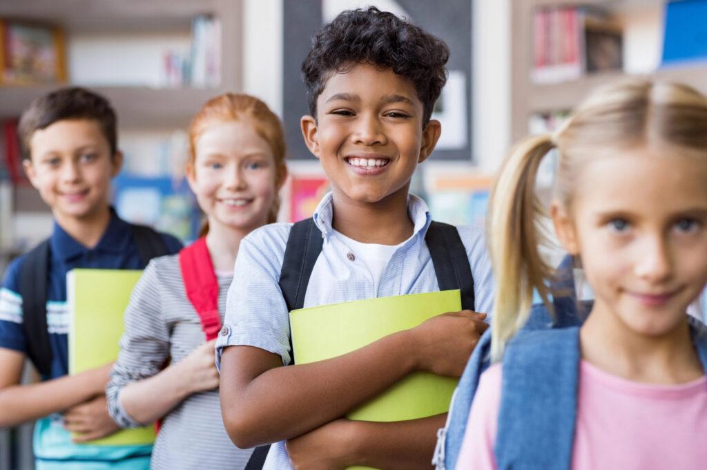 A diverse group of schoolchildren standing in a classroom, smiling at the camera with backpacks and colorful folders.