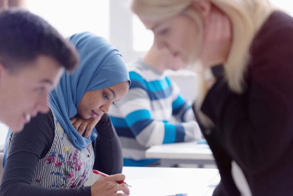 In a classroom, a girl wearing a blue hijab is writing at a desk while a blonde woman leans over to help.