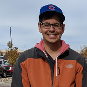 Smiling person wearing an orange and black jacket, a blue cap with a red C logo, and glasses, standing outdoors near a parking lot.