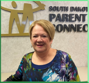 Smiling woman with short blonde hair wearing a colorful blouse, standing in front of the South Dakota Parent Connection sign.