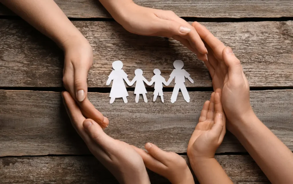 Several hands form a circular protective frame around a small paper cutout of a family of four on a wooden surface.