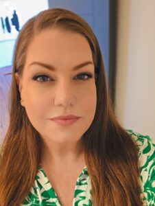 Close-up portrait of a woman with long brown hair wearing a green patterned shirt, looking at the camera.