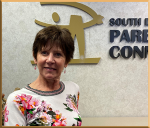 A woman with short brown hair wearing a floral blouse stands indoors in front of a wall with a gold logo and partial text.