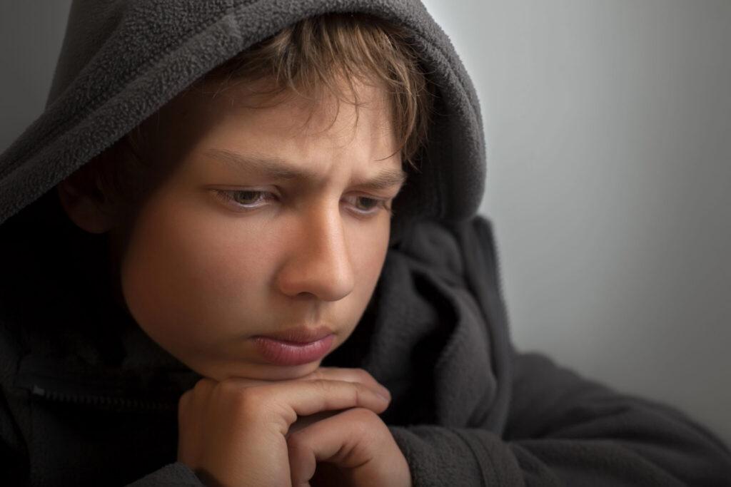 A young person wearing a dark hoodie rests their chin on clasped hands and looks down thoughtfully.
