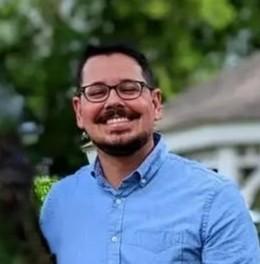 Smiling man wearing a light blue shirt and glasses standing outdoors with greenery and a house in the background.