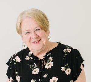 Smiling older woman with short blonde hair wearing a black floral blouse against a light background.