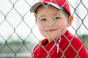 A young boy wearing a red cap and shirt smiles softly as he peers through a chain-link fence.