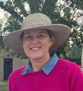 A smiling woman wearing a wide-brimmed straw hat and a pink sweater stands outdoors.
