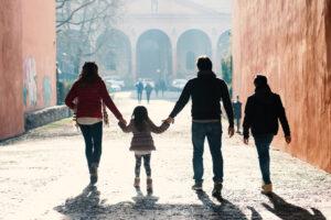 A family of four walks hand in hand down a sunlit cobblestone street between orange walls toward an arched gateway.