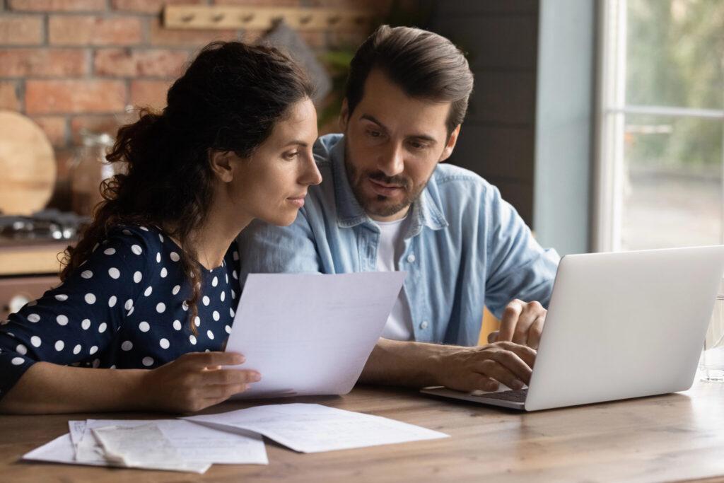 A man and a woman sit at a wooden table, reviewing papers with a laptop nearby.