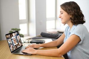 A woman in a light blue shirt sits at a wooden desk using a laptop for a video conference, with a grid of participants visible on the screen.