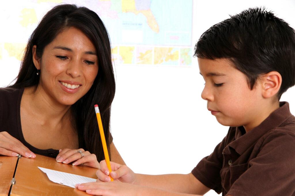 A woman guides a young boy as he writes with a pencil at a table.