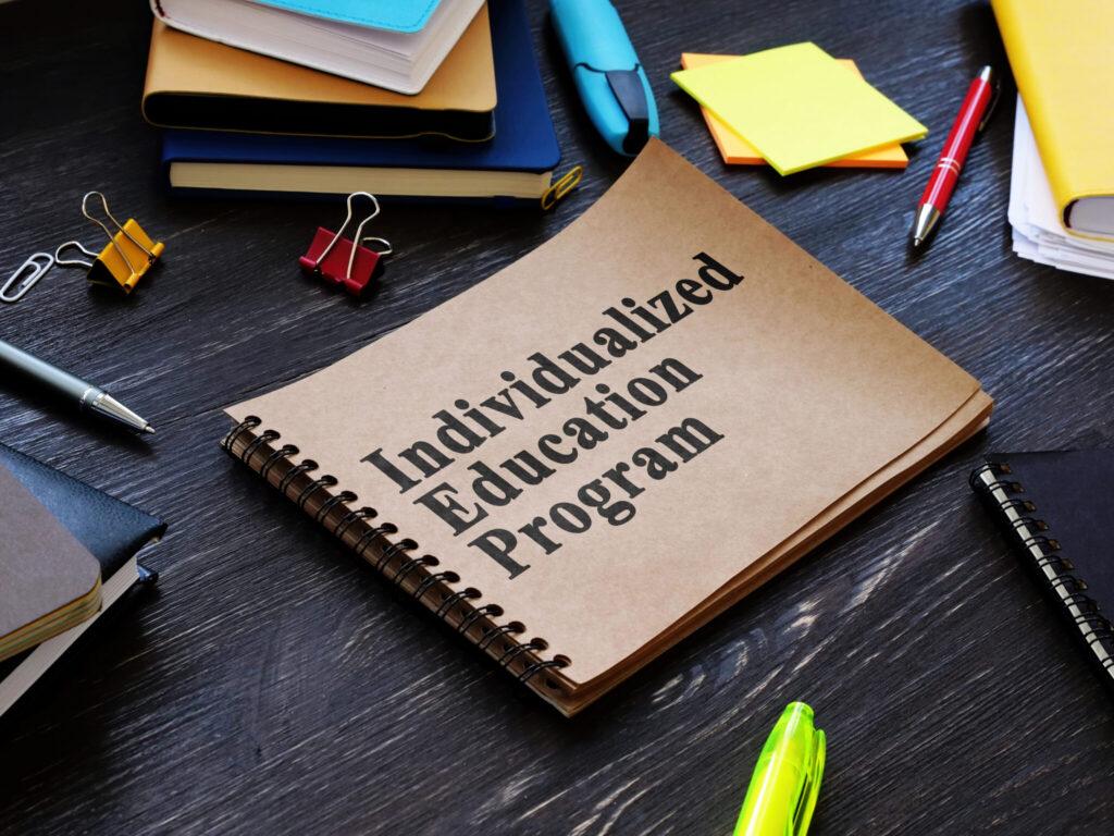 A spiral-bound notebook labeled “Individualized Education Program” sits on a dark desk surrounded by pens, clips, and sticky notes.